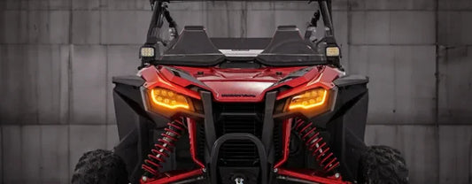 Front view of a red Honda Talon off-road vehicle, highlighting its aggressive design with bright amber headlights and suspension details, showcasing the vehicle's rugged appearance against a neutral background.