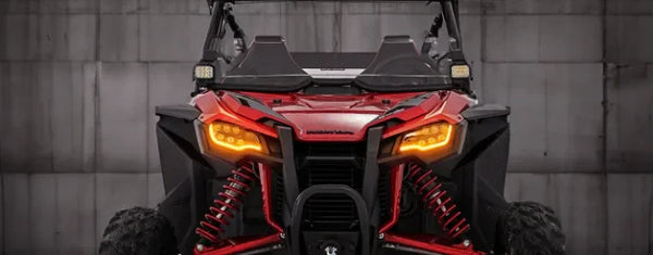 Front view of a red Honda Talon off-road vehicle, highlighting its aggressive design with bright amber headlights and suspension details, showcasing the vehicle's rugged appearance against a neutral background.